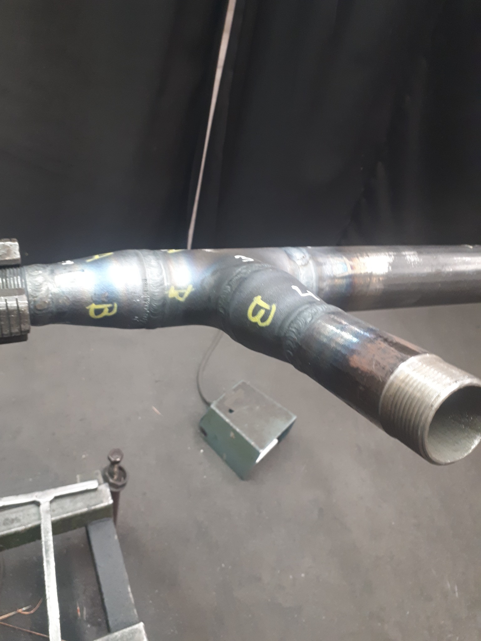 Spawany trójnik rurowy z oznaczeniami spoin na tle warsztatu, widoczne ślady obróbki i gwint na jednym z końców.