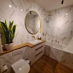 Nowoczesna łazienka z białymi płytkami imitującymi marmur, drewnianą podłogą i złotymi akcentami armatury oraz okrągłym lustrem, doświetlona punktowym oświetleniem sufitowym.