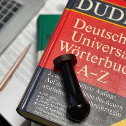 Słownik Duden leżący na biurku obok laptopa i ołówka, z czarnym stemplem na wierzchu.