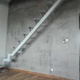 Surowe, szare wykończenie ściany imitujące beton, z drewnianą podłogą i minimalistycznymi, metalowymi schodami w Koszalinie. Widoczne gniazdka elektryczne na ścianie.