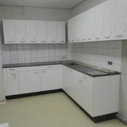 Nowoczesna, biała zabudowa kuchenna z szarym blatem i białymi płytkami na ścianie, widoczne niedociągnięcia wykończeniowe, takie jak listwa na blacie i niezabezpieczone gniazdka.