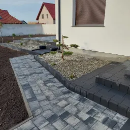 Nowoczesna aranżacja ogrodu z szarej kostki brukowej, schodów i rabat z kamykami oraz młodymi drzewkami iglastymi, widok na biały dom z brązowymi roletami.