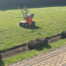 Pomarańczowa maszyna do rozkładania trawy z rolki stoi na trawniku obok wykopanego rowka, w którym leżą rolki trawy, w tle żywopłot.