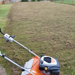 Pomarańczowa wertykulator spalinowy Stihl na trawniku po wertykulacji, widoczne resztki usuniętego filcu i mchu, w tle ogrodzenie i drzewa.