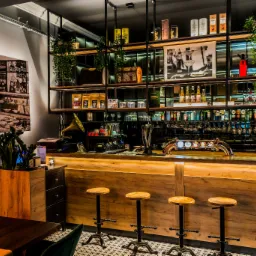 Wnętrze restauracji z drewnianym barem, hokerami na metalowych nogach, półkami z alkoholem i dekoracjami, gramofonem oraz czarno-białym zdjęciem na ścianie.