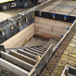 Wykop fundamentowy z deskowaniem i zbrojeniem, widoczny betonowóz w tle, ceglana ściana piwnicy z zamocowaniami.