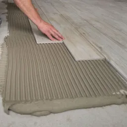 Układanie jasnych płytek imitujących drewno na podłodze, widoczne świeżo nałożone klej i dłoń dociskająca płytkę.