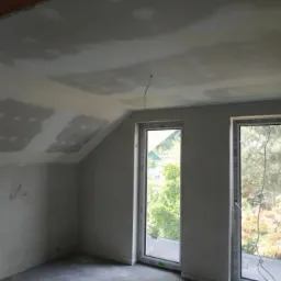 Pokój na poddaszu w trakcie wykańczania: ściany i skośny sufit pokryte gładzią gipsową, widoczne szpachlowane łączenia płyt, dwa okna z widokiem na zieleń na zewnątrz.