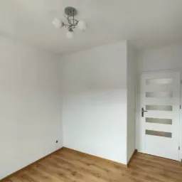 Pusty pokój po remoncie z drewnianą podłogą, białe ściany, białe drzwi z przeszkleniami i lampa sufitowa z czterema kloszami.