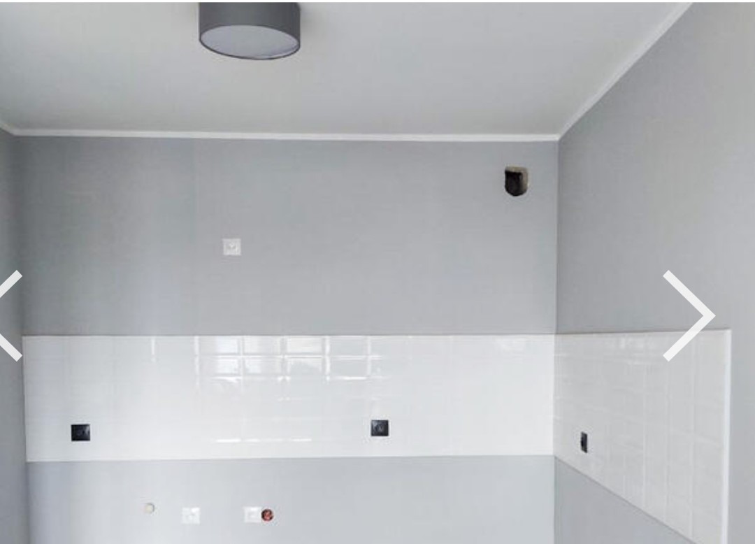 Fragment ściany w łazience z ułożonymi białymi, błyszczącymi płytkami w formie cegiełek, szara ściana powyżej z widocznymi otworami na instalacje, szare ściany boczne, sufit z szarą lampą.