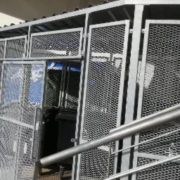 Metalowa wiata z siatki na pojemniki na odpady, częściowo otwarta, z widocznymi niebieskimi pojemnikami w środku, ustawiona obok schodów z metalową poręczą.