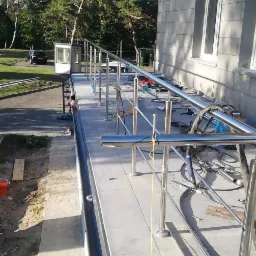 Montaż balustrady nierdzewnej przy budynku, widoczne narzędzia i elementy konstrukcyjne, otoczenie zieleni.