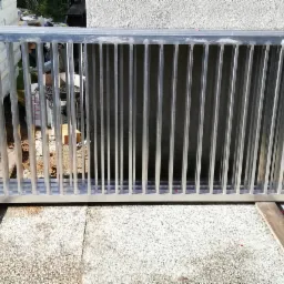 Srebrna, metalowa konstrukcja z pionowymi prętami, prawdopodobnie element bramy lub ogrodzenia, oparta na drewnianych legarach, widoczna sterta piasku i styropianu w tle.