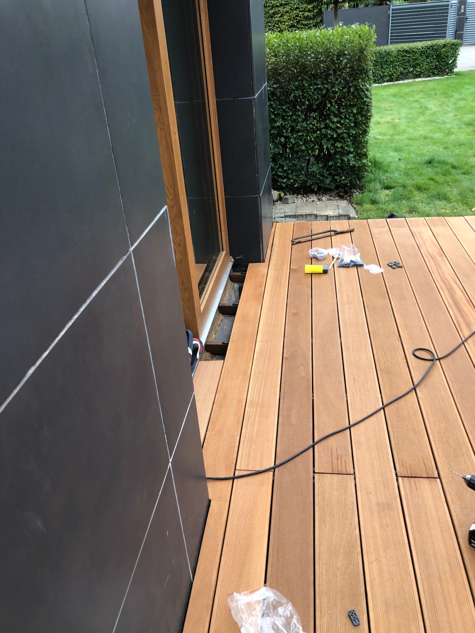 Montaż drewnianego tarasu przy nowoczesnym domu z czarną elewacją. Widoczne deski, narzędzia i elementy montażowe. Częściowy widok trawnika w tle.