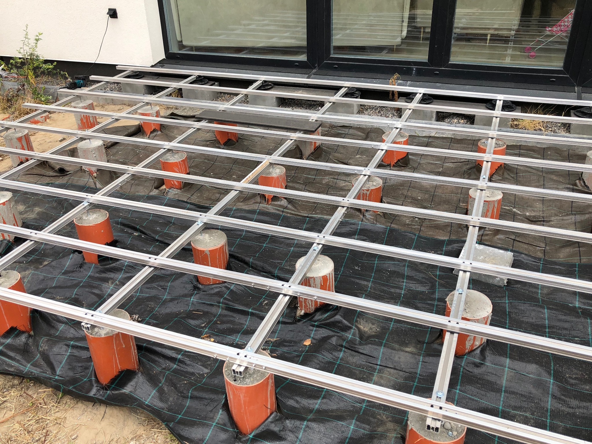 Konstrukcja tarasu z aluminiowych profili na betonowych słupkach z rur PCV, widoczna czarna mata geowłókninowa. Detal montażu, świeży beton.