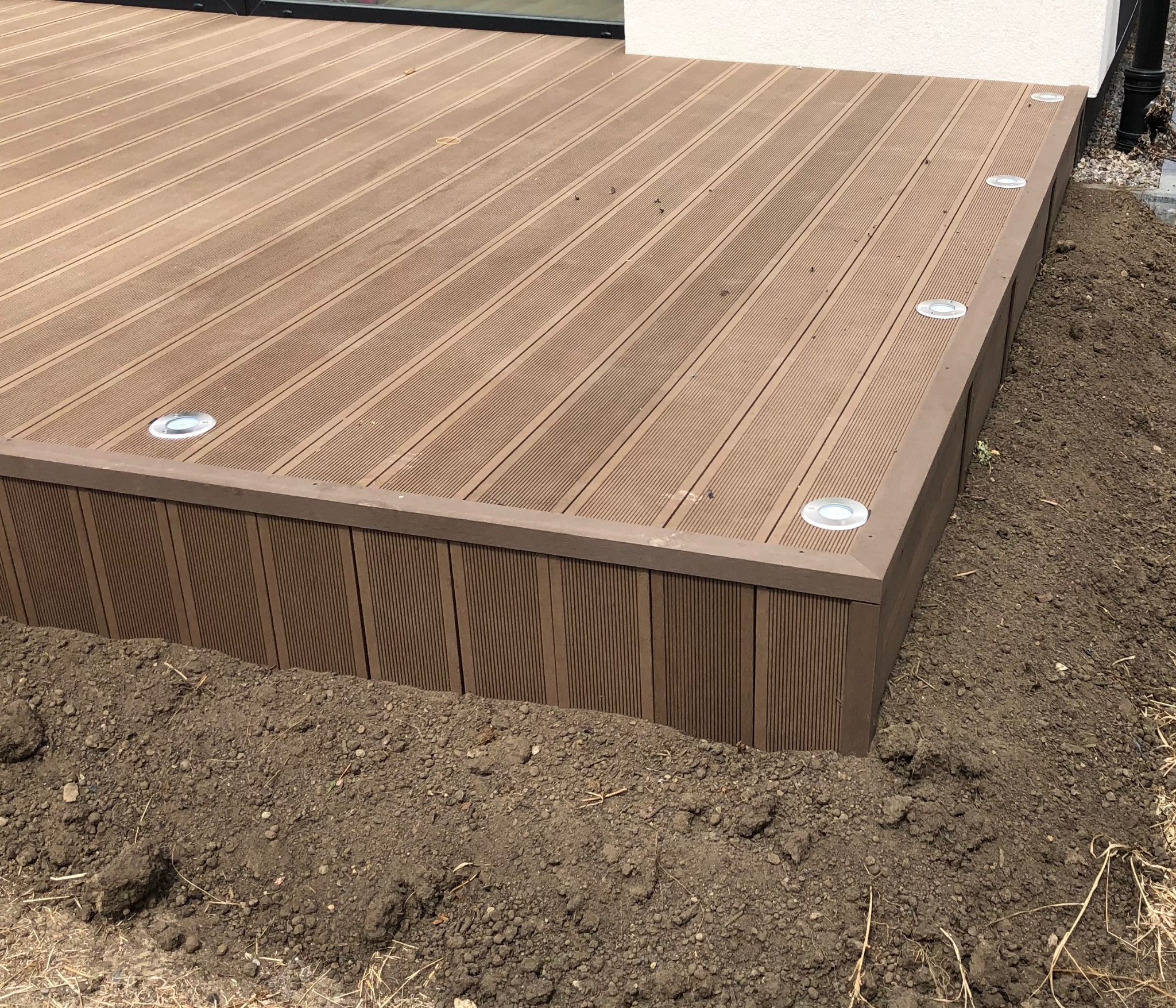 Nowoczesny taras z deski kompozytowej w odcieniach brązu, z wbudowanymi punktami świetlnymi, otoczony ziemią, częściowo widoczny budynek w tle.