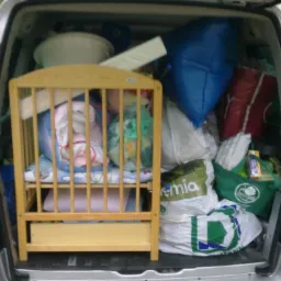 Załadowany bagażnik srebrnego vana z widocznym drewnianym łóżeczkiem dziecięcym, torbami z zakupami i innymi przedmiotami podczas przeprowadzki.