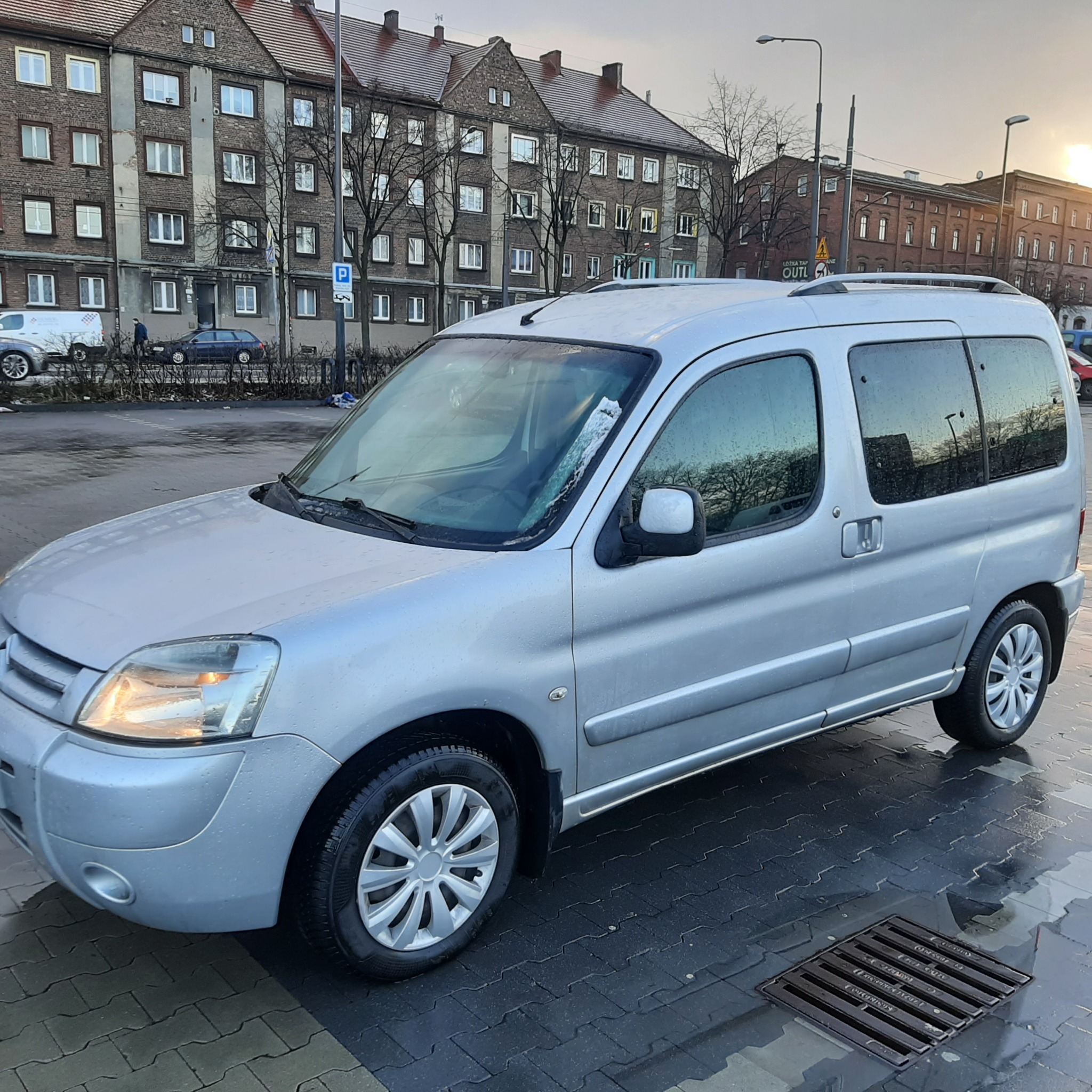 Srebrny bus Citroen Berlingo z relingami dachowymi, zaparkowany na mokrej kostce brukowej na tle budynków mieszkalnych i biurowych w pochmurny dzień.