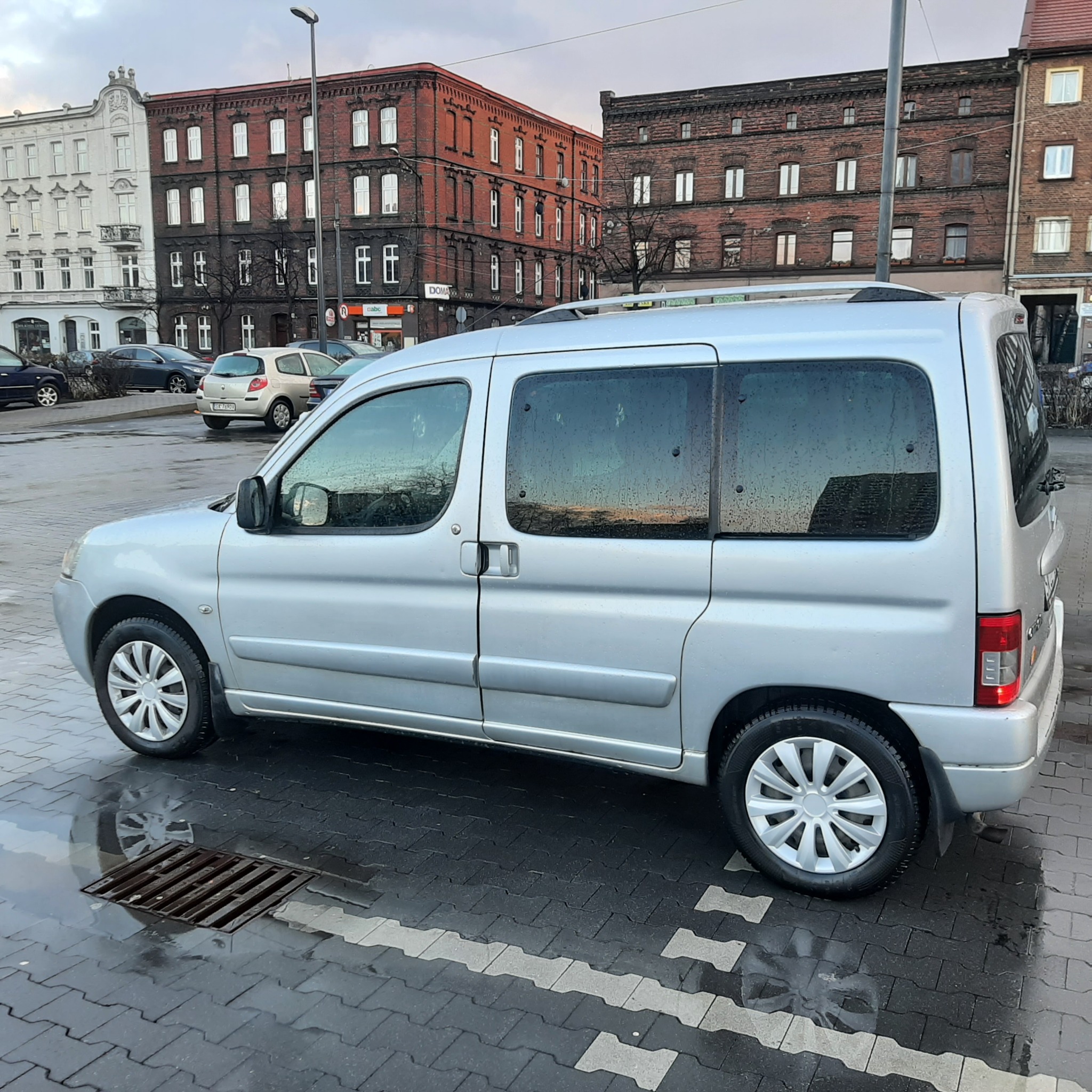 Srebrny bus zaparkowany na mokrym bruku z widokiem na kamienice w tle, widoczne krople deszczu na szybach.