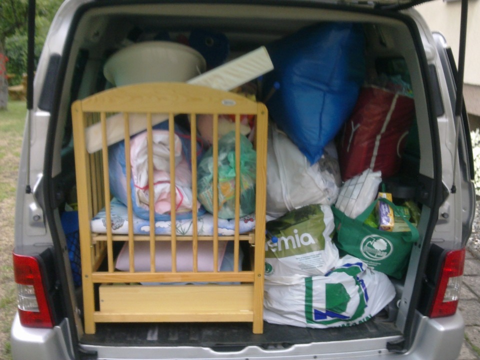 Załadowany bagażnik srebrnego vana z widocznym drewnianym łóżeczkiem dziecięcym, torbami z zakupami i innymi przedmiotami podczas przeprowadzki.