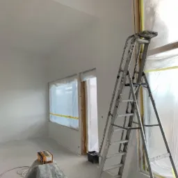 Wnętrze w trakcie prac wykończeniowych: ściany po nałożeniu gładzi, okna zabezpieczone folią i taśmą, drabina oparta o ścianę, podłoga zabezpieczona folią malarską, widoczny karton i wiadro.