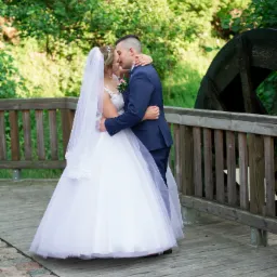 Para młoda w objęciach na drewnianym moście z fragmentem starego koła młyńskiego w tle.