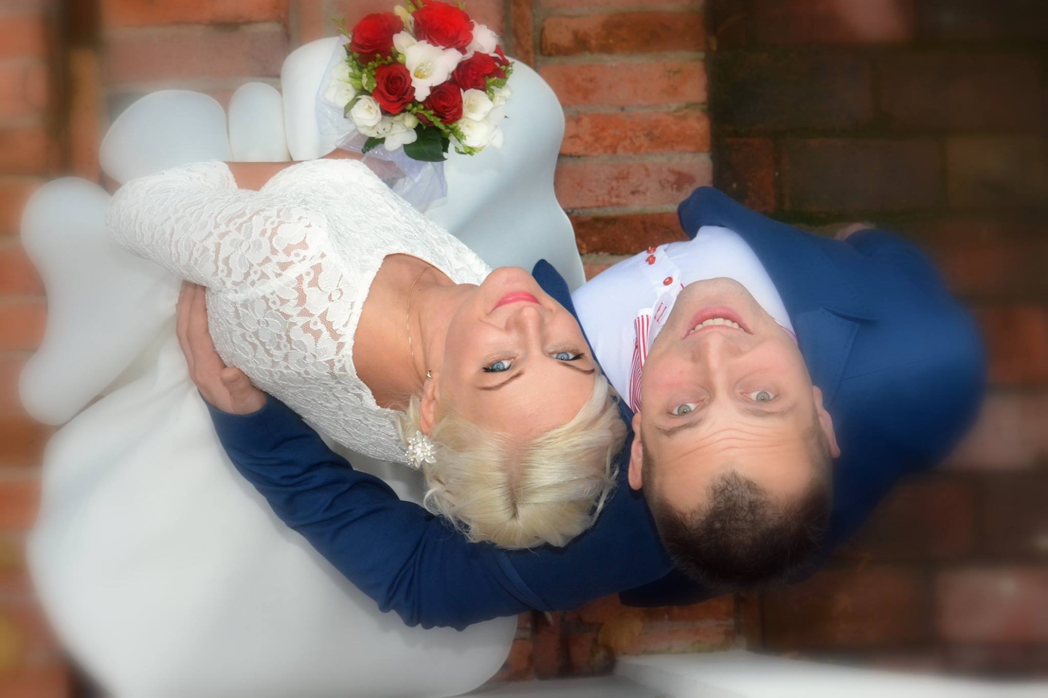 Para młoda, leżąca na białym krześle, trzymająca się za ręce, z bukietem czerwono-białych róż, sfotografowana z góry na tle ceglanej ściany.