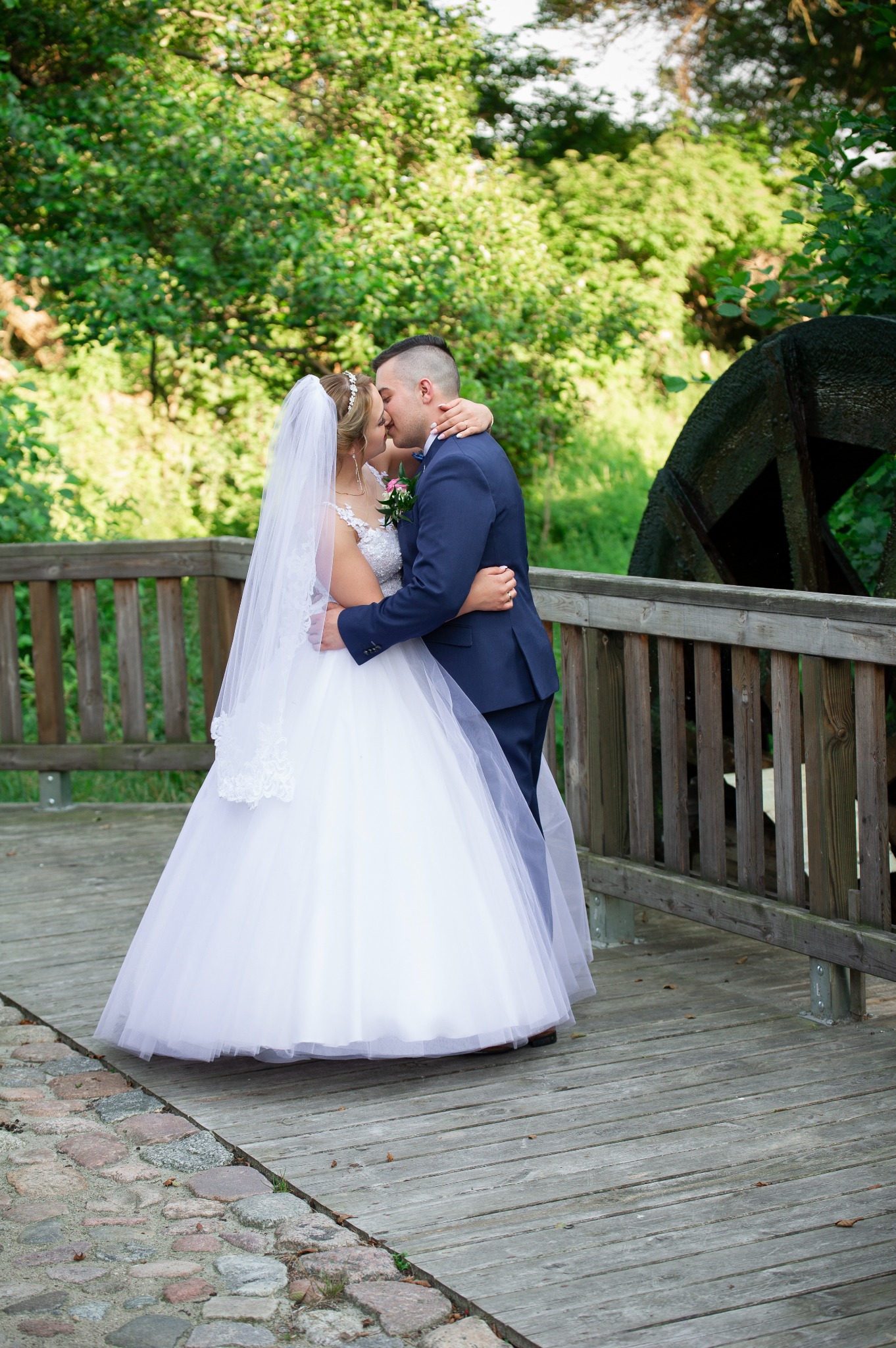 Para młoda w objęciach na drewnianym moście z fragmentem starego koła młyńskiego w tle.