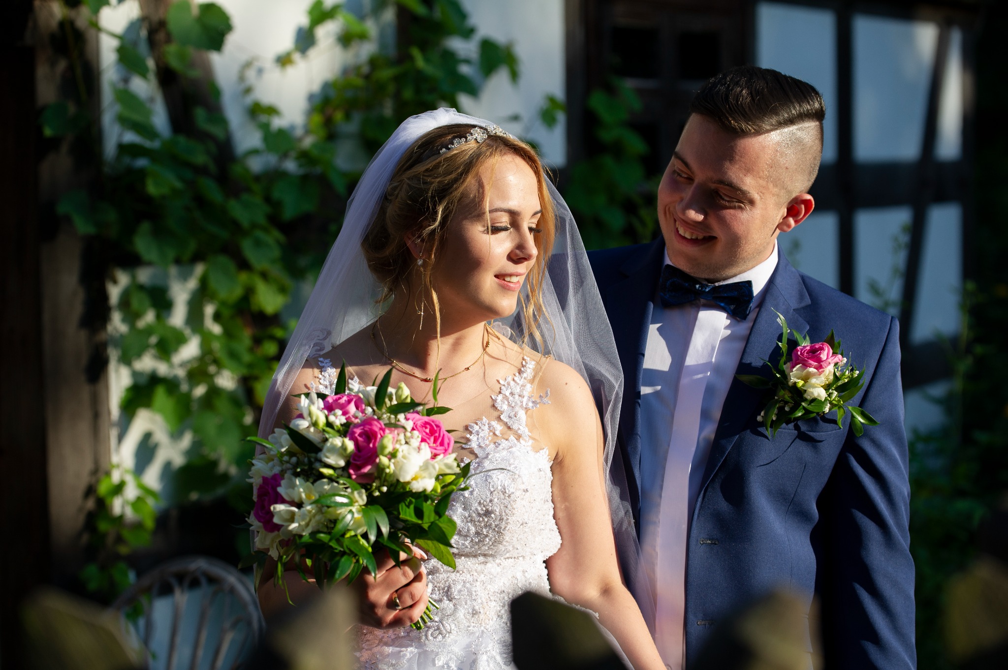 Uśmiechnięta panna młoda w koronie i welonie trzyma bukiet różowych i białych kwiatów, obok niej pan młody w granatowym garniturze z butonierką, oboje stoją na tle zieleni i białej ściany.