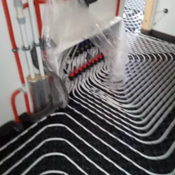 Układ rur grzewczych w jodełkę, czarne podłoże, białe rury, rozdzielacz zakryty folią, czerwone i białe rury pionowe przy ścianie.