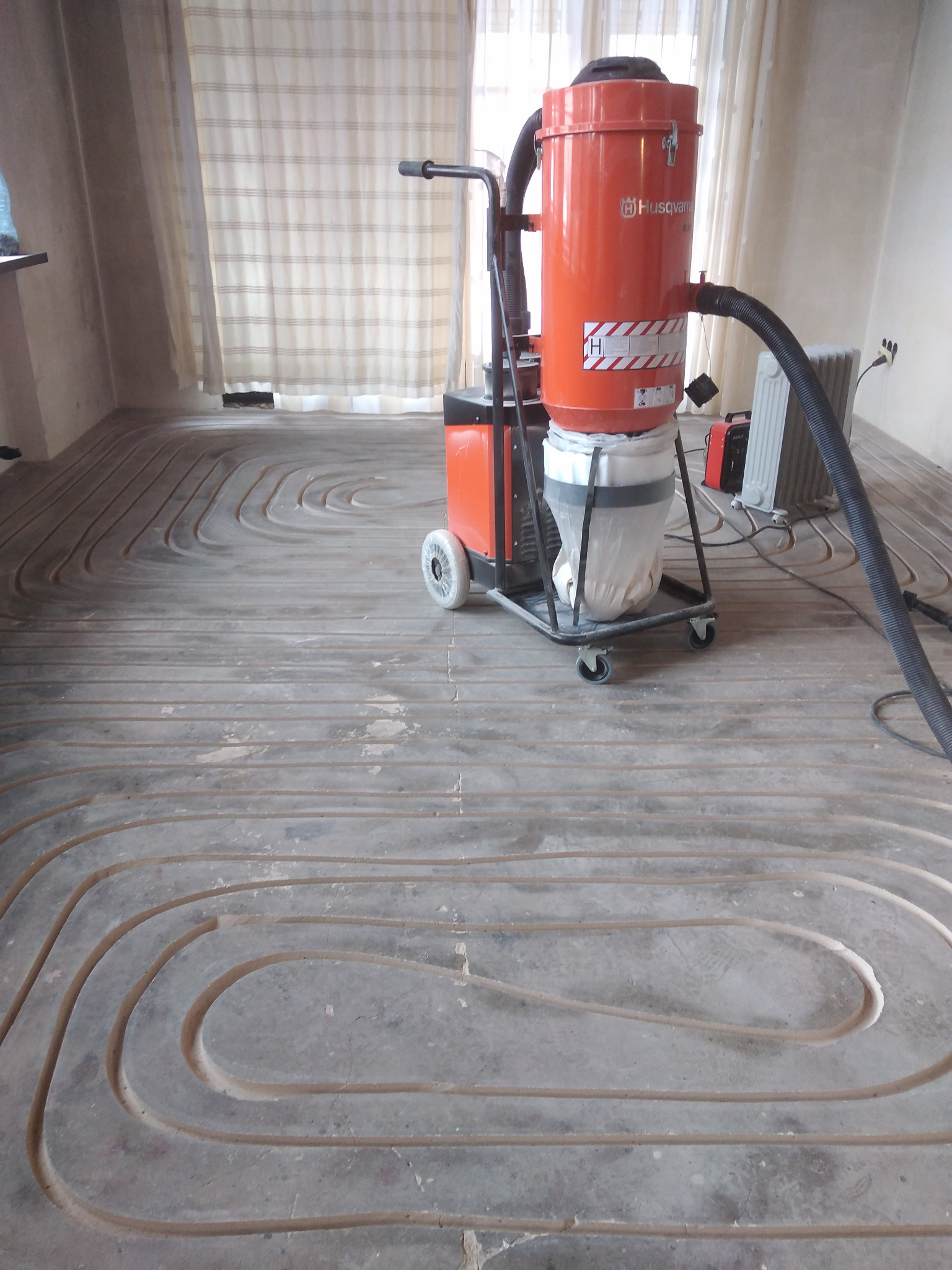 Wyfrezowane rowki w podłodze pod instalację ogrzewania podłogowego, widoczny odkurzacz przemysłowy Husqvarna na kółkach, przygotowanie do montażu rur grzewczych.