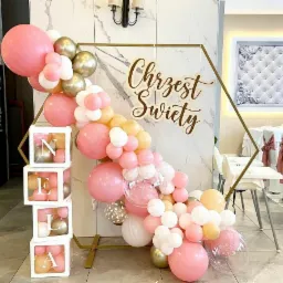 Dekoracja balonowa w odcieniach różu, bieli i złota z napisem 'Chrzest Święty' i imieniem 'NELA' w przezroczystych kostkach, ustawiona na tle marmurowej ściany w sali bankietowej.
