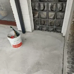 Świeżo nałożona, jasnoszara warstwa wyrównująca podłoże, widoczna przy ścianie z luksferami, obok stoi wiadro z narzędziami i resztkami materiału budowlanego.