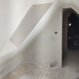 LECH-POL Leszek Duks Usługi Ogólnobudowlane - Poddasze po świeżym pomalowaniu na biało, widoczne skośne ściany z oknem dachowym, gniazdko elektryczne wystające ze ściany i podłoga z resztkami farby, otwór drzwiowy prowadzący do ciemnego...
