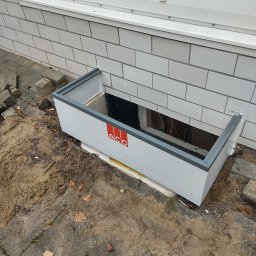 LECH-POL Leszek Duks Usługi Ogólnobudowlane - Montaż okna piwnicznego z osadnikiem ACO, widoczne kable wewnątrz, mur z białej cegły i fragment chodnika z piaskiem.
