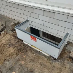 Montaż okna piwnicznego z osadnikiem ACO, widoczne kable wewnątrz, mur z białej cegły i fragment chodnika z piaskiem.