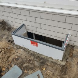 LECH-POL Leszek Duks Usługi Ogólnobudowlane - Montaż okna piwnicznego z obudową ACO, widoczne detale montażowe oraz logo producenta na białej obudowie, ceglany mur i fragment okna na górze.
