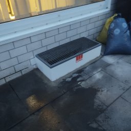 LECH-POL Leszek Duks Usługi Ogólnobudowlane - Okolice okna z odpływem liniowym marki ACO, widoczne cegły elewacyjne, worki na śmieci w tle, mokra kostka brukowa.