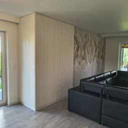 Nowoczesne wnętrze: panel ścienny z pionowymi listwami w jasnym odcieniu, tapeta z motywem liści i szara podłoga imitująca drewno. Częściowo widoczny czarny narożnik.