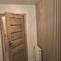 Nowoczesne wnętrze: drewniane lamele na ścianie, otwarta dębowa drzwi z poziomymi wstawkami, biały grzejnik i szara podłoga. Minimalistyczna lampa sufitowa.