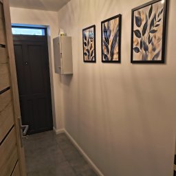 LECH-POL Leszek Duks Usługi Ogólnobudowlane - Korytarz po remoncie: szare płytki, białe ściany z trzema obrazami w czarnych ramkach, szara szafka i drzwi. Widoczna lampa sufitowa i fragment drewnianych drzwi.