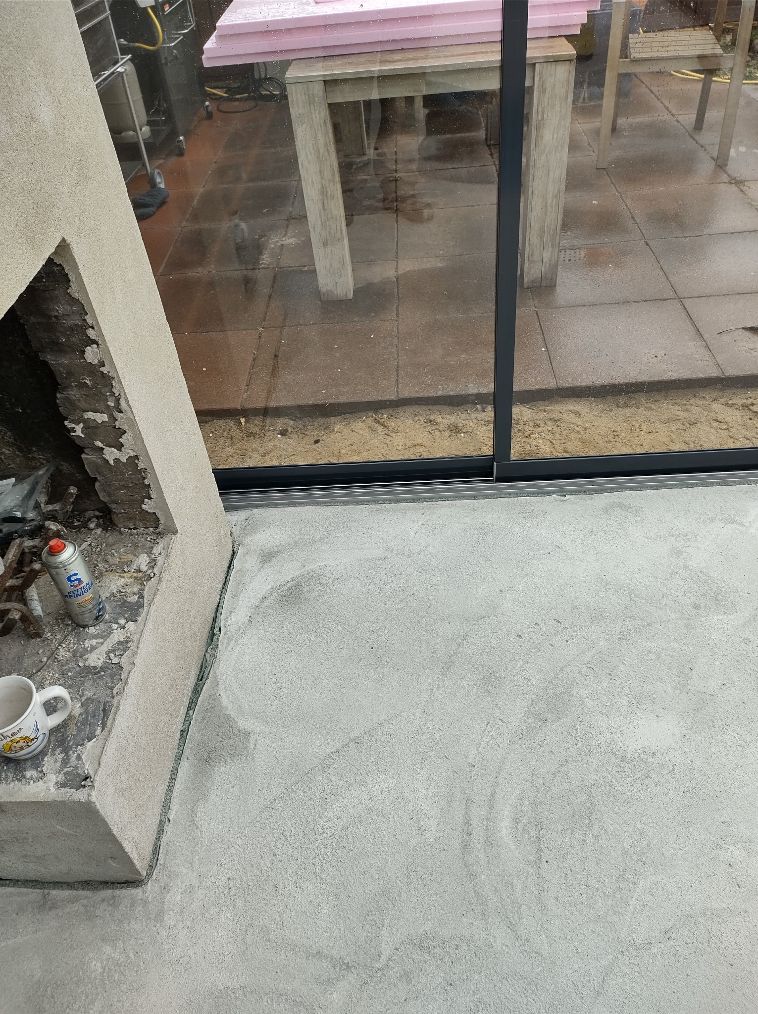 Świeżo wylana, jasnoszara wylewka podłogowa w pomieszczeniu z dużym oknem przesuwnym, widoczny fragment ściany z resztkami starego tynku, puszka pianki montażowej i kubek.