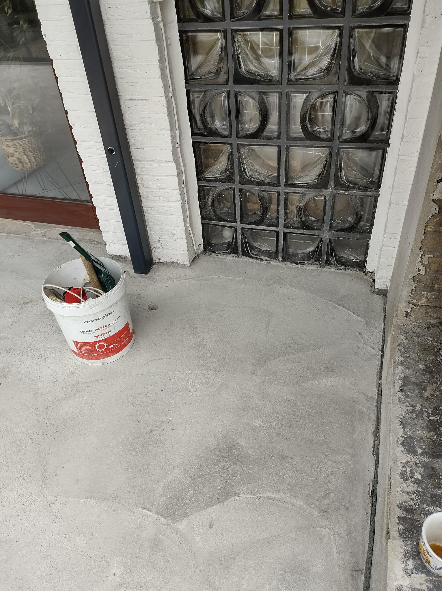 Świeżo nałożona, jasnoszara warstwa wyrównująca podłoże, widoczna przy ścianie z luksferami, obok stoi wiadro z narzędziami i resztkami materiału budowlanego.