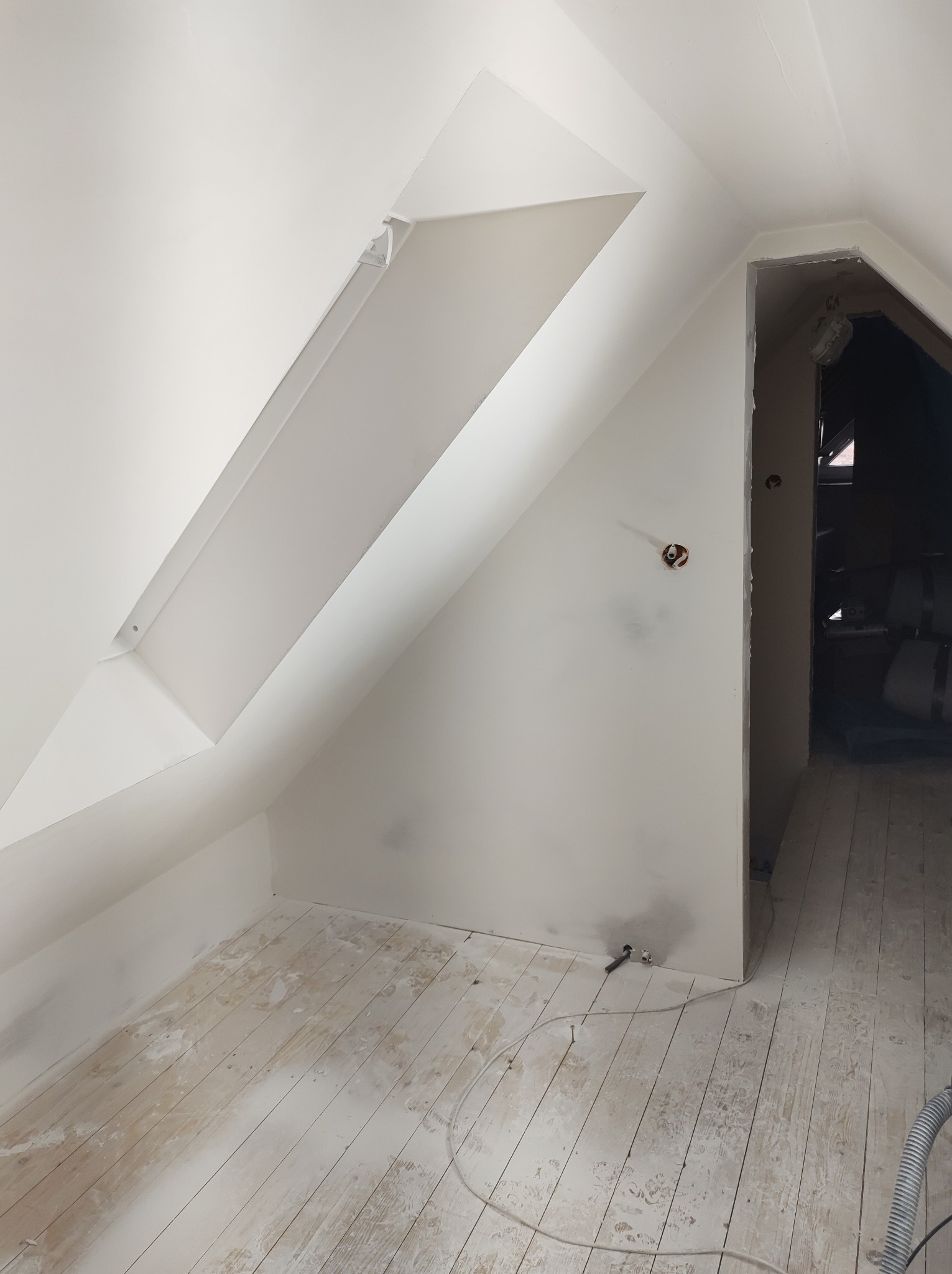 Poddasze po świeżym pomalowaniu na biało, widoczne skośne ściany z oknem dachowym, gniazdko elektryczne wystające ze ściany i podłoga z resztkami farby, otwór drzwiowy prowadzący do ciemnego...