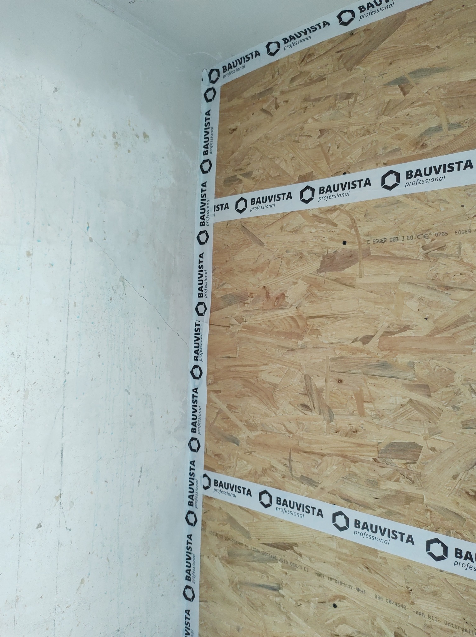 Montaż płyty OSB na ścianie, zabezpieczonej taśmą Bauvista Professional wzdłuż krawędzi, widoczna surowa ściana w rogu pomieszczenia.