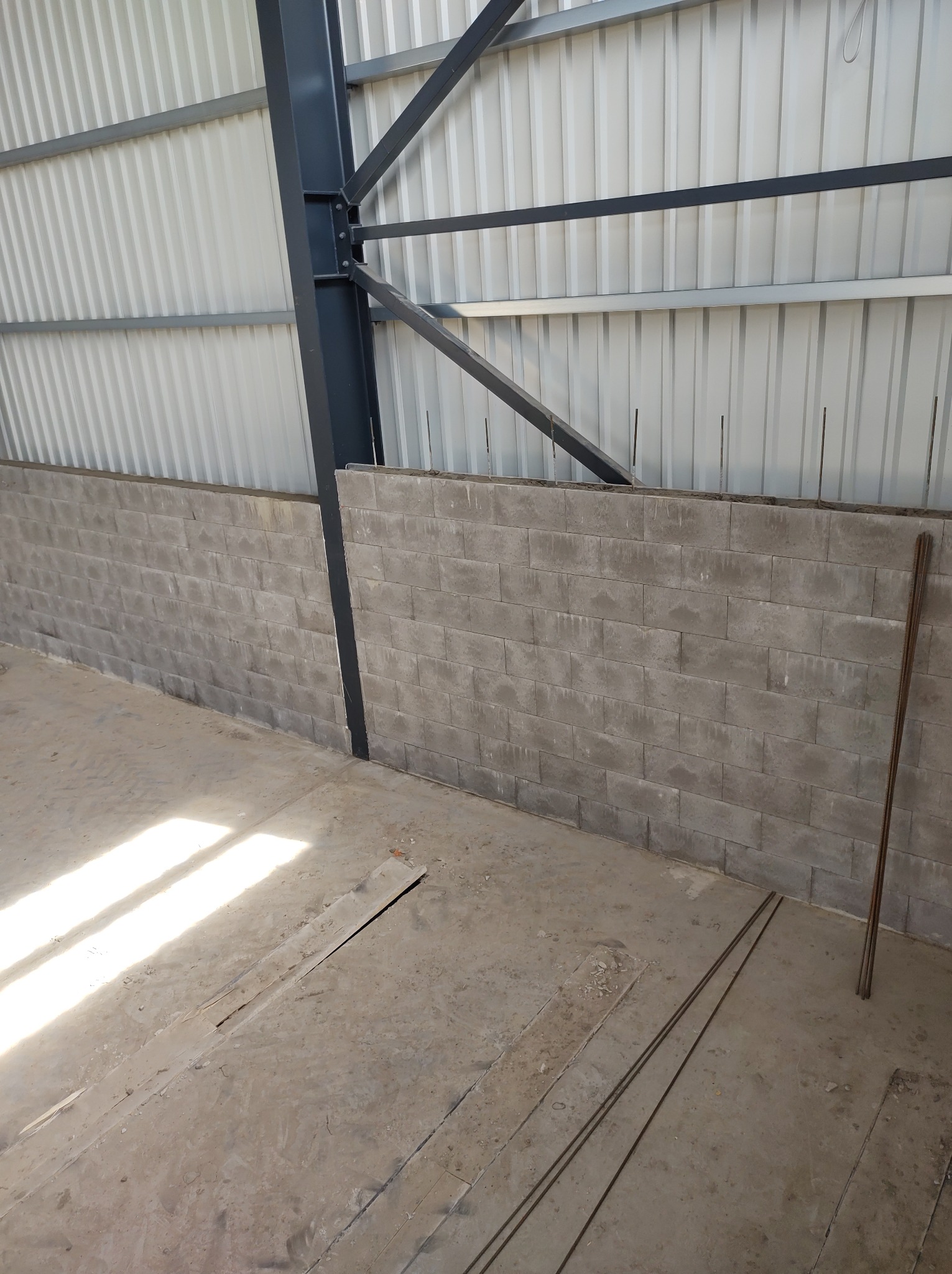 Wnętrze hali w trakcie budowy: murowana ściana z bloczków betonowych, widoczne pręty zbrojeniowe, konstrukcja stalowa dachu i posadzka w stanie surowym, jasne światło słoneczne wpadające do wnętrza.