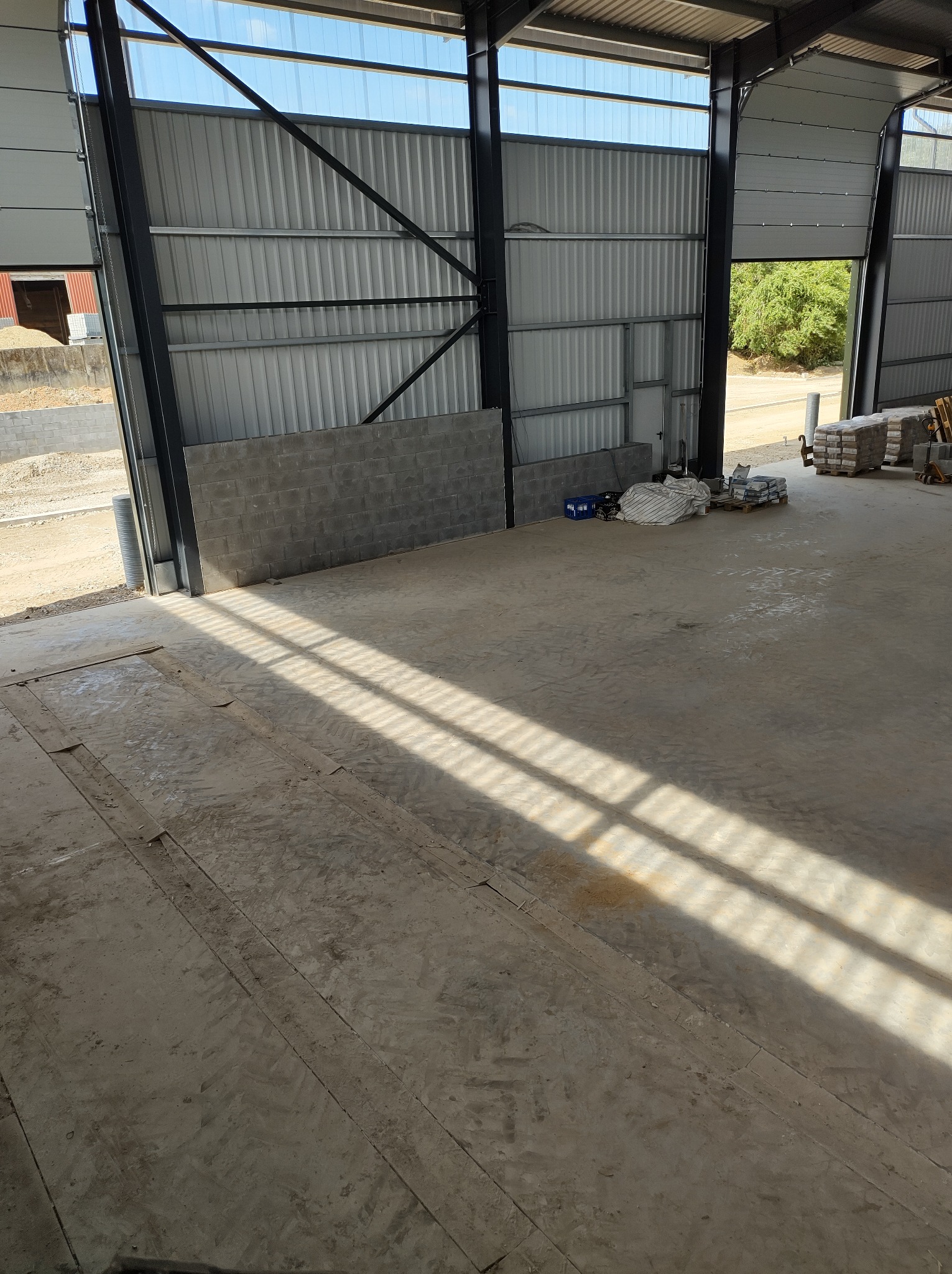 Wnętrze hali w trakcie prac budowlanych, widoczne materiały budowlane, betonowe podłoże z wyraźnymi śladami po maszynach i wpadające przez otwór w dachu promienie światła.