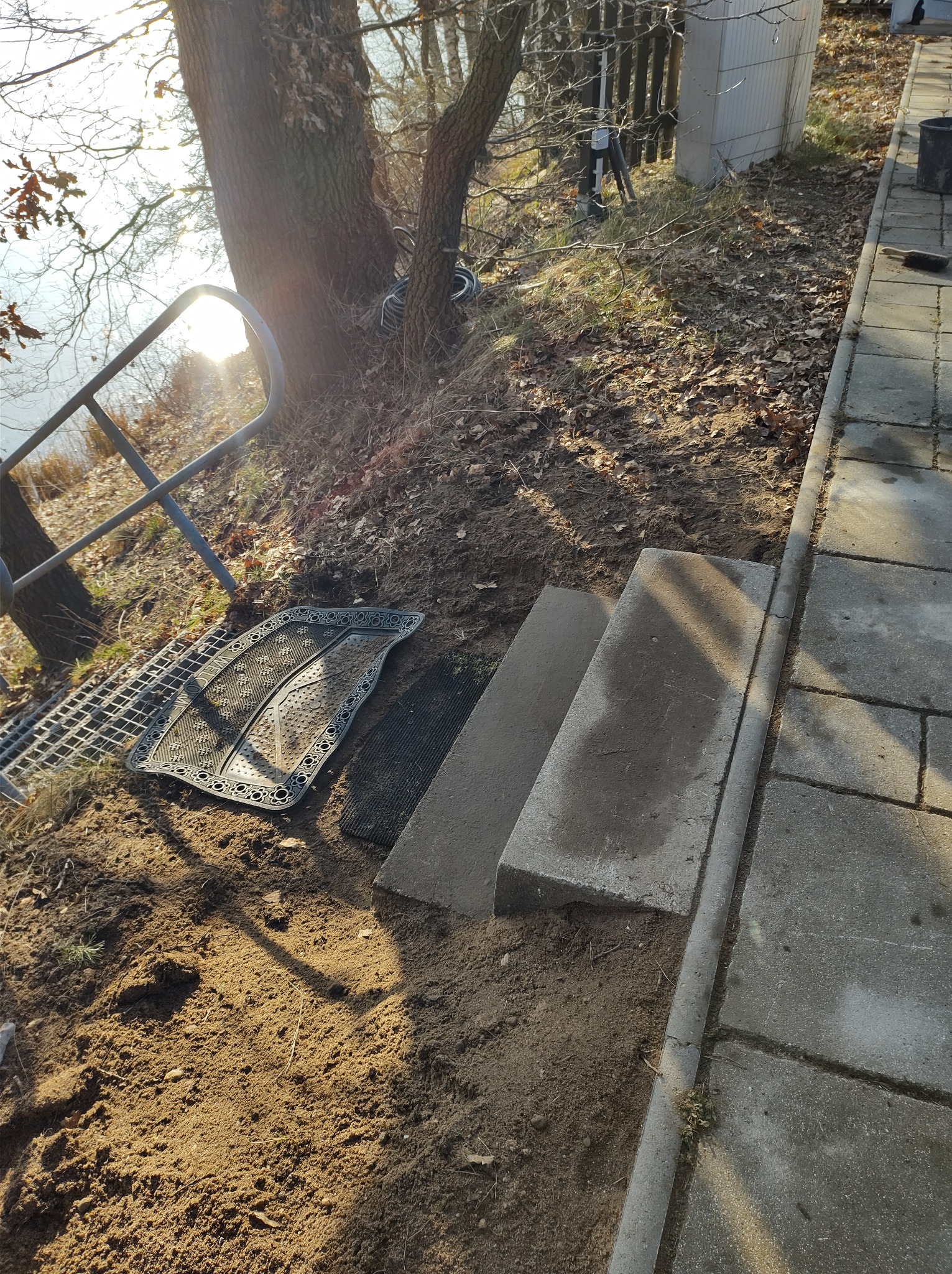 Montaż schodów zewnętrznych z betonowych płyt w ogrodzie przy jeziorze, widoczne przygotowane podłoże i fragment istniejącego chodnika.