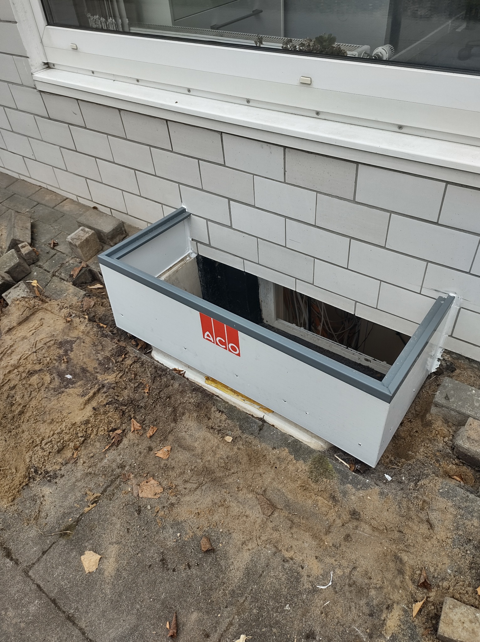 Montaż okna piwnicznego z osadnikiem ACO, widoczne kable wewnątrz, mur z białej cegły i fragment chodnika z piaskiem.