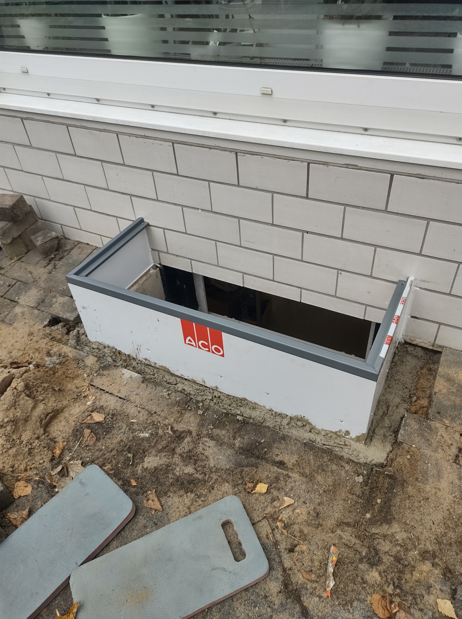 Montaż okna piwnicznego z obudową ACO, widoczne detale montażowe oraz logo producenta na białej obudowie, ceglany mur i fragment okna na górze.